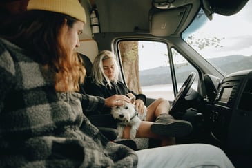A couple sitting in the front seats of their campervan with their West Highland White Terrier on their laps, using their campervan as a holiday home
