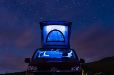 The VW Transporter stealthy pop top roof open and parked under a starry night glowing light inside illuminating the camper in a field.