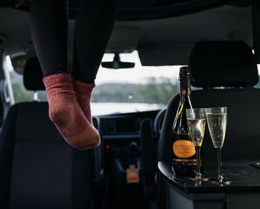 Person sitting on their pop top roof with legs dangling into the back of a Ford Transit Custom with two glasses of prosecco and a bottle next to their feet upgrading their summer.